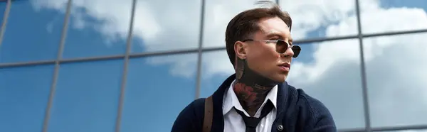 Handsome young man with tattoos relaxes outside a contemporary building under a clear sky. — Stock Photo