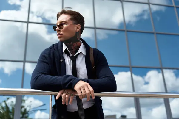 Young man with tattoos leans casually on a railing in front of a reflective building. — Stockfoto