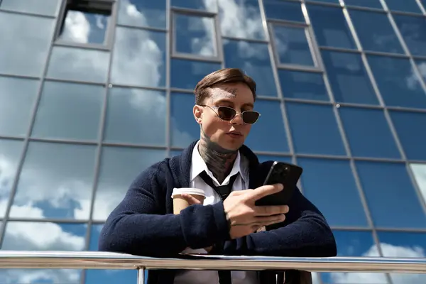Uomo con tatuaggi gode di caffè guardando il suo smartphone vicino a una struttura di vetro. — Foto stock