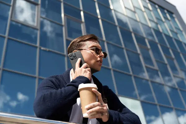 Handsome young man with tattoos talks on the phone while enjoying coffee outside a building. — Stock Photo