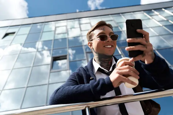 Handsome young man with tattoos smiles as he sips coffee and looks at his phone in the city. — Stock Photo