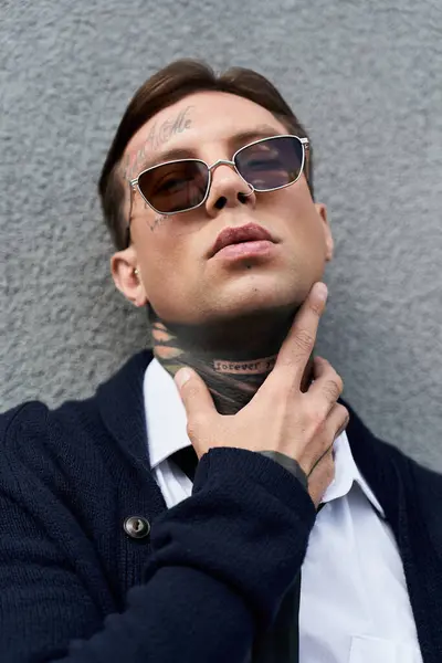 Man with tattoos shows a relaxed pose against a gray wall, sporting trendy sunglasses and attire. — Stock Photo
