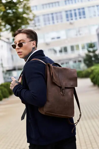 Handsome young man with tattoos strolls through a city area wearing sunglasses and a backpack. — Stock Photo