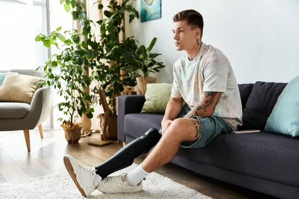 Handsome young man sits thoughtfully on a couch, showcasing his prosthetic leg in a bright space. — Stock Photo