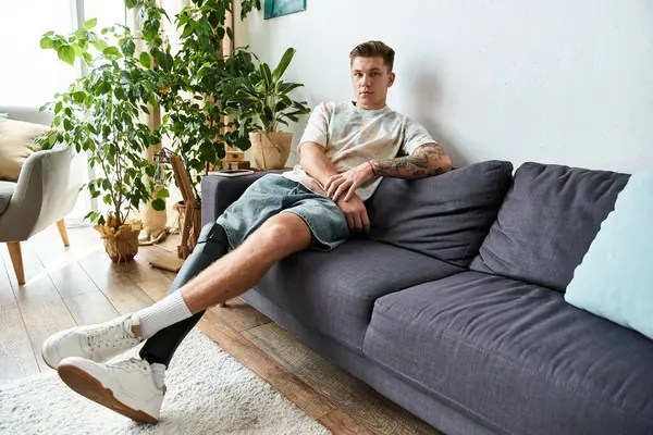 A young handsome man with a prosthetic leg relaxes on a comfortable sofa surrounded by plants. — Stock Photo
