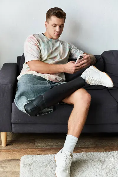 Handsome young man with a prosthetic leg relaxes on a modern couch, absorbed in his smartphone. — Stock Photo