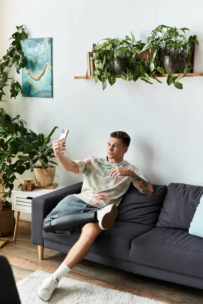 Sitting comfortably on a sofa, a handsome young man with a prosthetic leg takes a selfie, smiling. — Stock Photo