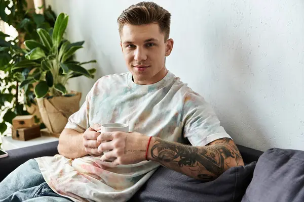 A cheerful young man sits on a sofa, sipping a warm drink. — Stock Photo