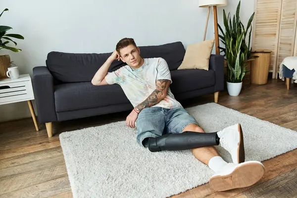 Schöner junger Mann sitzt auf einem weichen Teppich neben einer Couch und präsentiert sein prothetisches Bein bequem. — Stockfoto