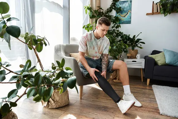 A young handsome man sits in a modern chair, adjusting his prosthetic leg among houseplants. — Stock Photo