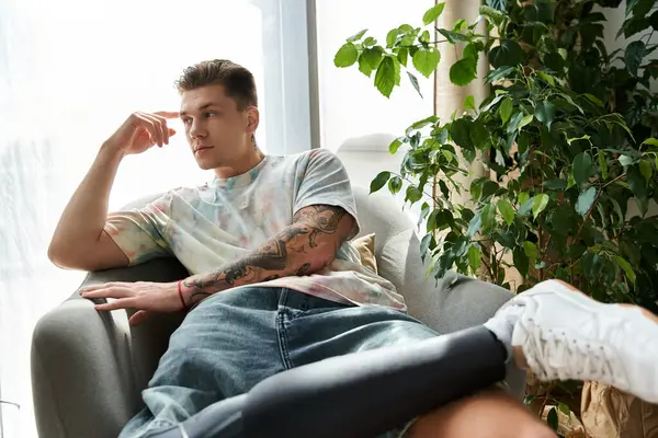 Young man thoughtfully resting on a cozy couch, showcasing his prosthetic leg and vibrant tattoos. — Stock Photo