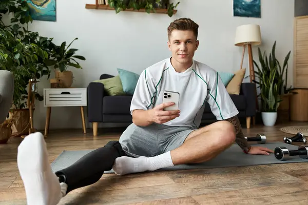 Joven hombre guapo se sienta en una alfombra con una pierna protésica, sonriendo mientras usa su teléfono inteligente. - foto de stock