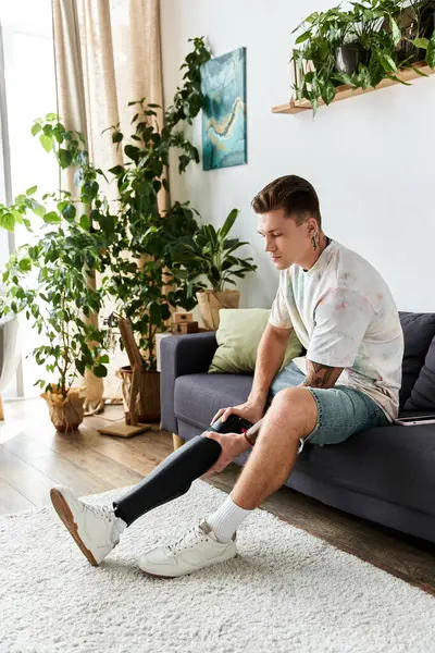 Young man sits on a cozy couch, thoughtfully adjusting his prosthetic leg in a sunny space. — Stock Photo