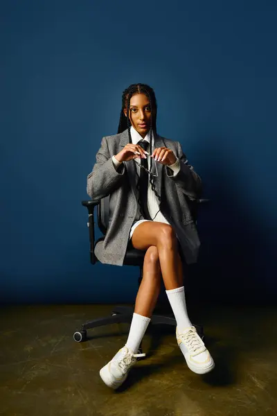 A confident young woman sits in a chic chair, flaunting her trendy outfit against a bold backdrop. — Stock Photo