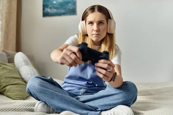 Una bella adolescente si siede a gambe incrociate sul suo letto, completamente concentrata sul gioco, indossando cuffie. — Foto stock