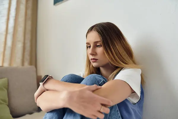 Una adolescente reflexiva se sienta en el suelo, abrazando sus rodillas en su elegante habitación. - foto de stock