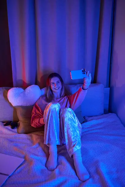 A teenage girl enjoys an evening in her stylish room, capturing a joyful selfie amidst soft lights. — Stock Photo