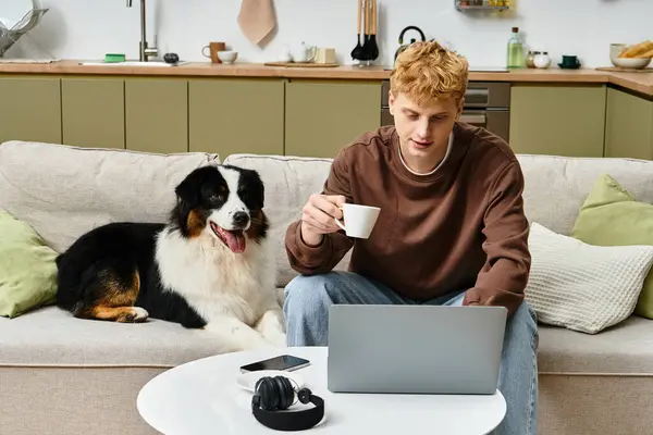 Jeune homme assis sur un canapé avec un berger australien tout en sirotant du café et en utilisant un ordinateur portable. — Photo de stock