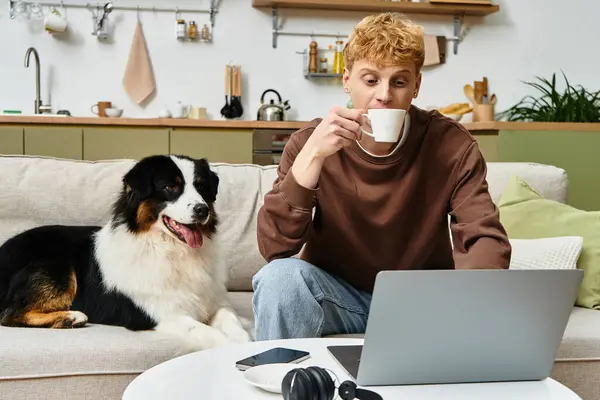 A young redhead man sips coffee while working on his laptop beside his Australian Shepherd dog. — Stock Photo