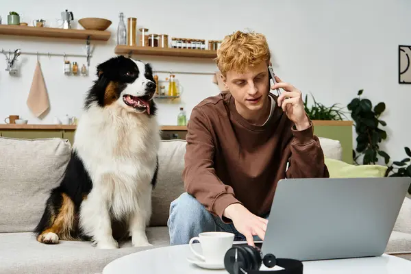 A young man with red hair interacts with his laptop while his dog sits beside him in a cozy home. — Stock Photo
