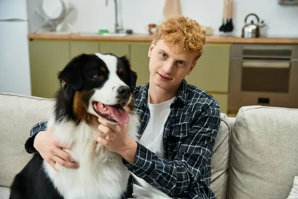 In a modern apartment, a young man with red hair plays with his cheerful Australian Shepherd. — Stock Photo