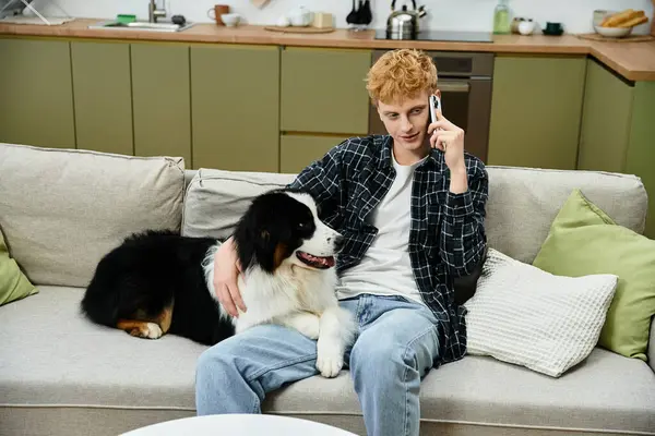 A young redhead relaxes on a couch, chatting on his phone while petting his Aussie. — Stock Photo