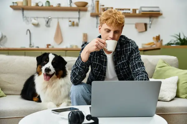 A young redhead man enjoys a warm drink while working on a laptop at home with his dog. — Stock Photo