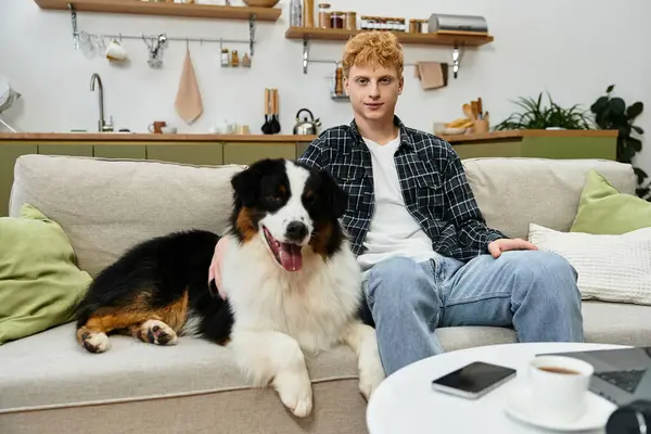 A young redhead man relaxed on the couch with his cheerful Australian Shepherd dog in a modern home. — Stock Photo