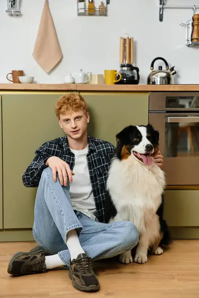 A redhead man sits on the floor of a stylish kitchen, smiling at his loyal dog beside him. — Stock Photo