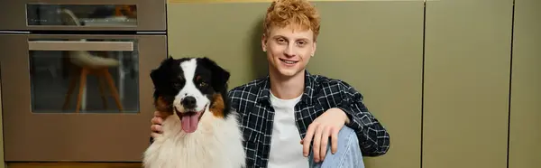 A young redhead man spends time with his cute Australian Shepherd in a modern apartment, banner — Stock Photo