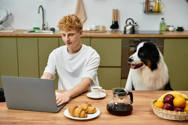 A young man with red hair focuses on his laptop while his dog sits beside him in the kitchen. — Stock Photo