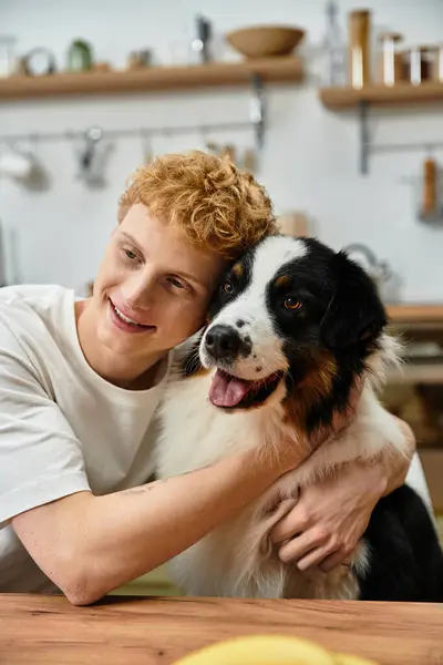 A cheerful young man with red hair embraces his adorable Australian Shepherd in a modern space. — Stock Photo
