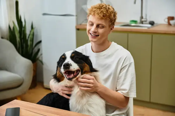 Young redhead man smiles while cuddling his cheerful dog at home. A cozy moment shared. — Stock Photo