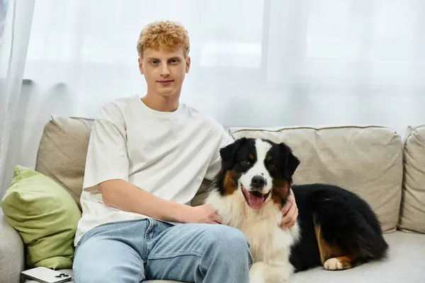 A red-haired young man relaxes on a sofa with his friendly Australian Shepherd dog. — Stock Photo