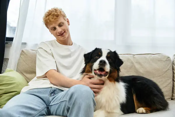 Un jeune rouquin se détend sur un canapé, caressant amoureusement son chien berger australien à la maison. — Photo de stock