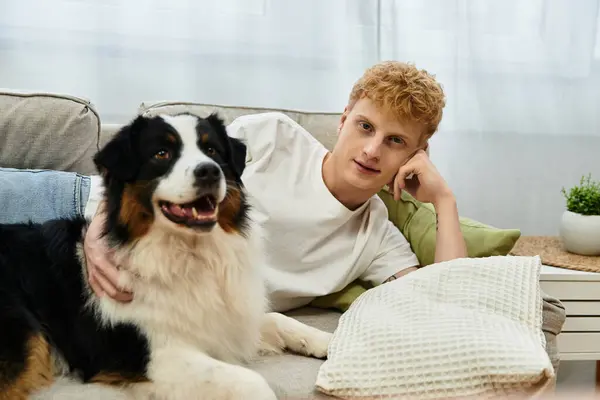 A young redhead relaxes on a couch with his cheerful Australian Shepherd in a modern apartment. — Stock Photo