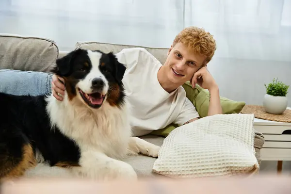 A cheerful young man with red hair relaxes on the couch with his adorable Australian Shepherd dog. — Stock Photo