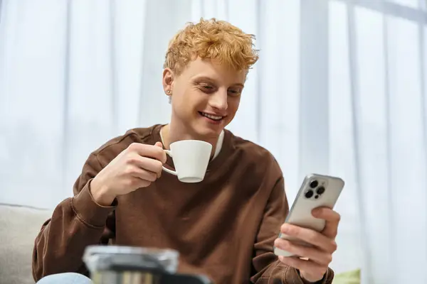A young man with red hair sips coffee and smiles at his phone in a modern living space. — Stock Photo