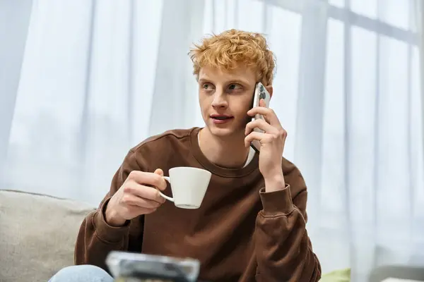 A young redhead man enjoys a cup while talking on the phone in his cozy apartment. — Stock Photo