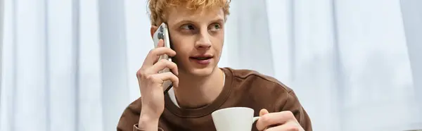 A handsome young man with red hair talks on his phone while sipping coffee at home. — Stock Photo