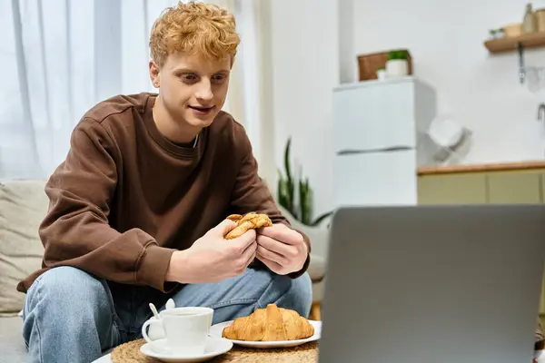 Un giovane uomo con i capelli rossi ricci gode di un croissant e caffè sul divano, incentrato sul suo computer portatile. — Foto stock