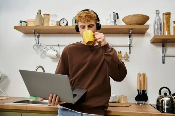 Young man with red hair focuses on his laptop while sipping coffee in a chic kitchen. — Stock Photo