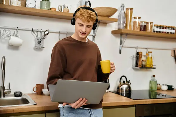 A young man with red hair engages with his laptop while holding a warm drink in a stylish kitchen. — Stock Photo