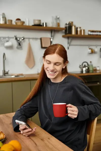 Charming young man with long hair sips coffee in his stylish apartment, checking his phone. — Stock Photo