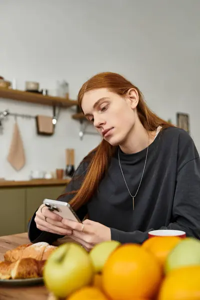 A stylish young man with long hair relaxes at home, checking his phone and enjoying breakfast. — Stock Photo