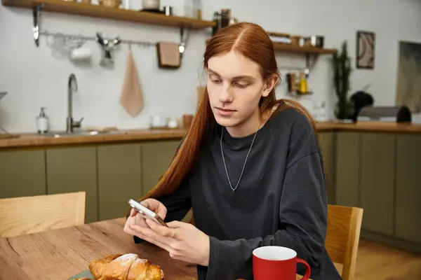 In a modern apartment, a young man relaxes at breakfast, checking his phone — Stock Photo