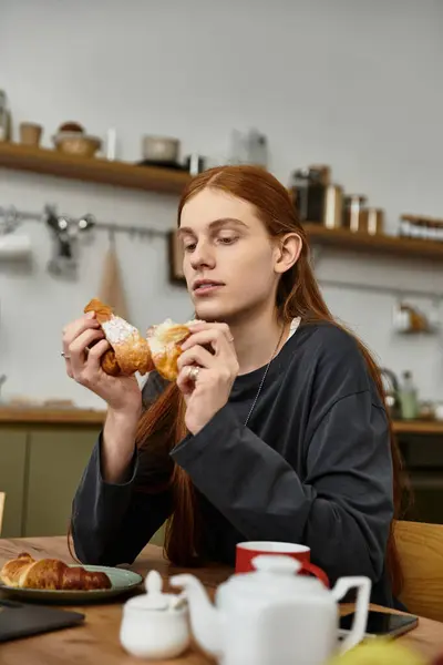 A young man with long hair savors a delicious pastry in a warm and inviting kitchen. — Stock Photo