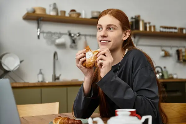 Ein stilvoller junger Mann genießt ein Croissant an einem gemütlichen Küchentisch und reflektiert seinen Morgen. — Stockfoto