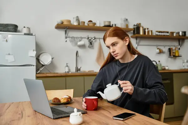 Un joven elegante con el pelo largo vierte el té en una taza roja mientras se relaja en casa. - foto de stock