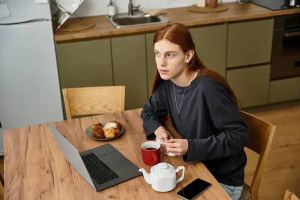 A young guy with long hair sits at a stylish table, sipping tea and enjoying pastries in his home. — Stock Photo
